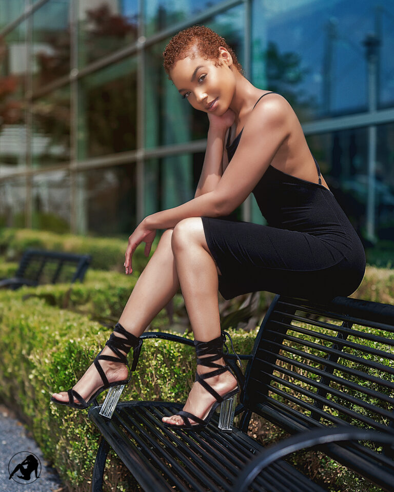 Model in a black dress seated on a bench in front of a glass building